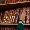 A person reaching for a red encyclopedia on a neatly organized bookshelf in a library.
